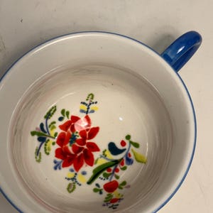 May include: A white ceramic bowl with a blue rim and handle. The interior features a vibrant floral design with red, yellow, and blue accents. The bowl is designed for serving soups, cereals, or other dishes.