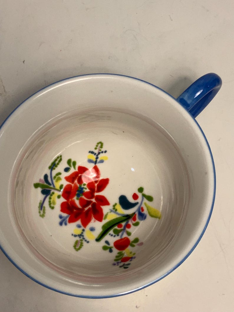 May include: A white ceramic bowl with a blue rim and handle. The interior features a vibrant floral design with red, yellow, and blue accents. The bowl is designed for serving soups, cereals, or other dishes.