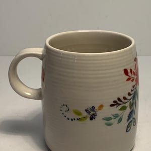 Puede incluir: Taza de cer&aacute;mica color crema con dise&ntilde;o floral. La taza tiene un asa curva y un cuerpo cil&iacute;ndrico con estr&iacute;as horizontales. El dise&ntilde;o floral incluye detalles en rojo, azul y verde. Capacidad aproximada de 350 ml.