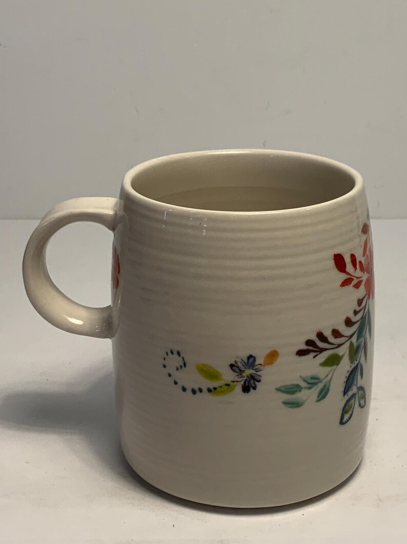 Puede incluir: Taza de cer&aacute;mica color crema con dise&ntilde;o floral. La taza tiene un asa curva y un cuerpo cil&iacute;ndrico con estr&iacute;as horizontales. El dise&ntilde;o floral incluye detalles en rojo, azul y verde. Capacidad aproximada de 350 ml.