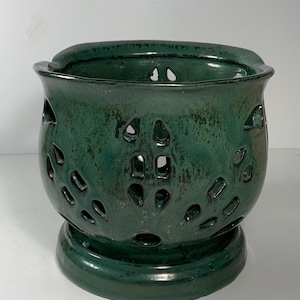 May include: A dark green ceramic candle holder with a round base and a bowl-shaped top. The sides of the holder feature decorative cut-out patterns. The candle holder has a glossy finish and is designed to hold a candle.