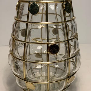 Op de afbeelding: Een heldere glazen vaas met een bolvormige vorm, omhuld door een goudkleurige metalen kooi. De kooi heeft een rasterachtig ontwerp met cirkelvormige accenten. De vaas is ontworpen om bloemen of andere decoratieve items in te bewaren.