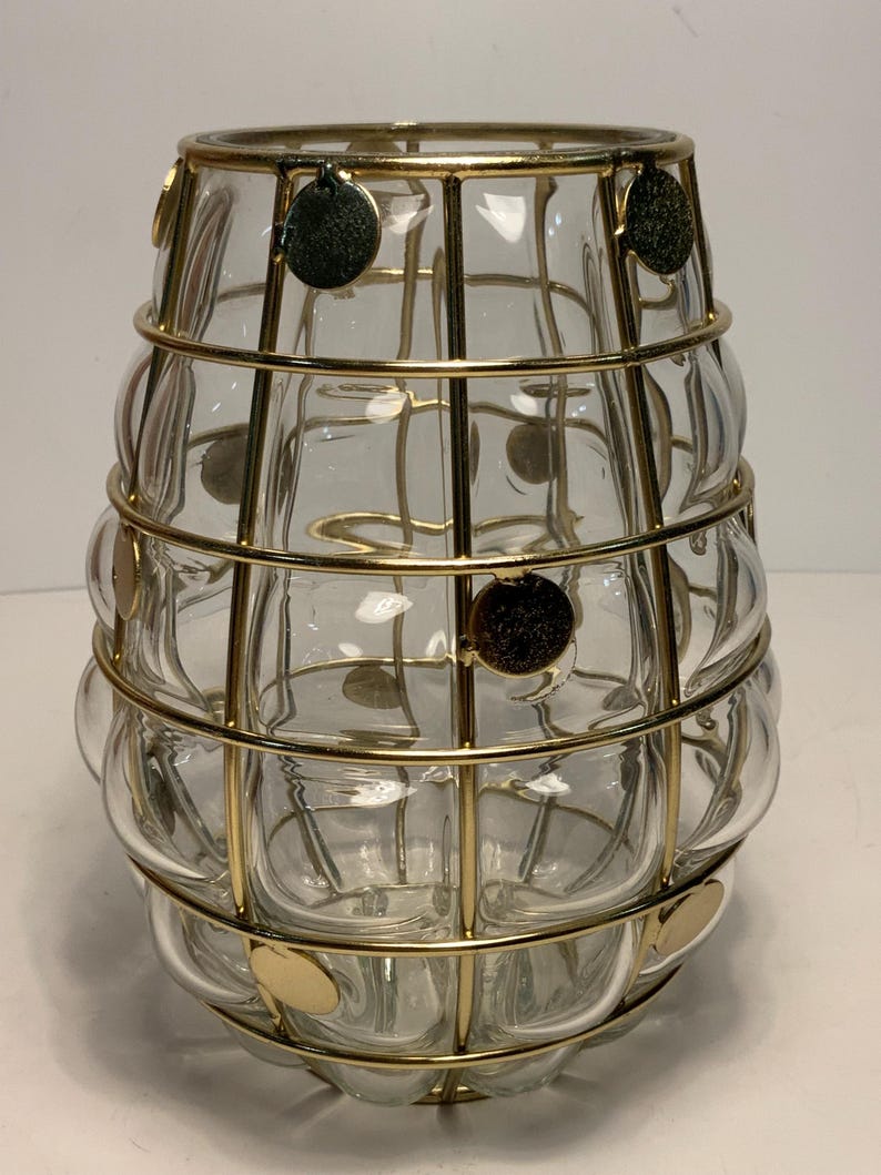 Op de afbeelding: Een heldere glazen vaas met een bolvormige vorm, omhuld door een goudkleurige metalen kooi. De kooi heeft een rasterachtig ontwerp met cirkelvormige accenten. De vaas is ontworpen om bloemen of andere decoratieve items in te bewaren.