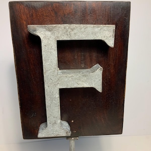 May include: A weathered metal letter "F" is mounted on a dark brown wooden plaque. The letter has a distressed, industrial appearance, and the wood has a rich, aged look. The sign is attached to a metal pole, suggesting it's a sign or display piece.