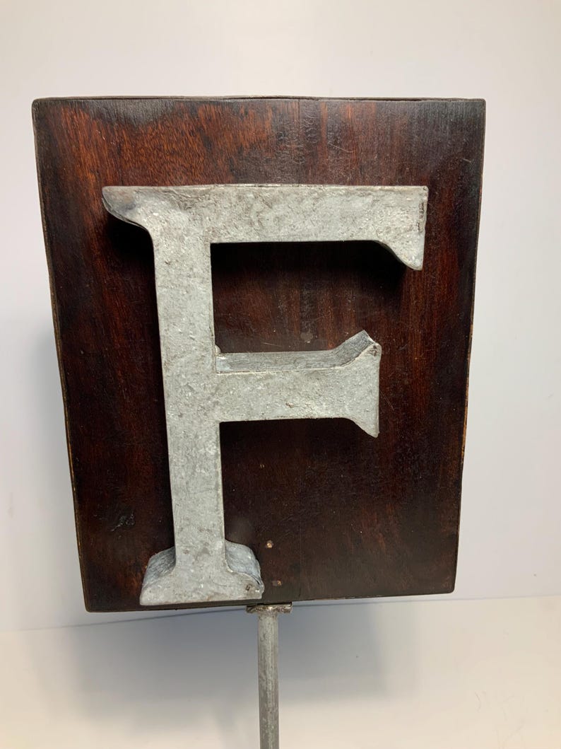 May include: A weathered metal letter "F" is mounted on a dark brown wooden plaque. The letter has a distressed, industrial appearance, and the wood has a rich, aged look. The sign is attached to a metal pole, suggesting it's a sign or display piece.
