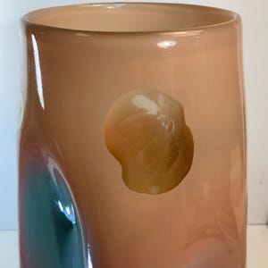 May include: A tall, cylindrical vase with a peach-colored exterior. The glass has a glossy finish with abstract shapes of teal and brown. The vase has a wide opening and a smooth, rounded base. The design is modern and artistic.