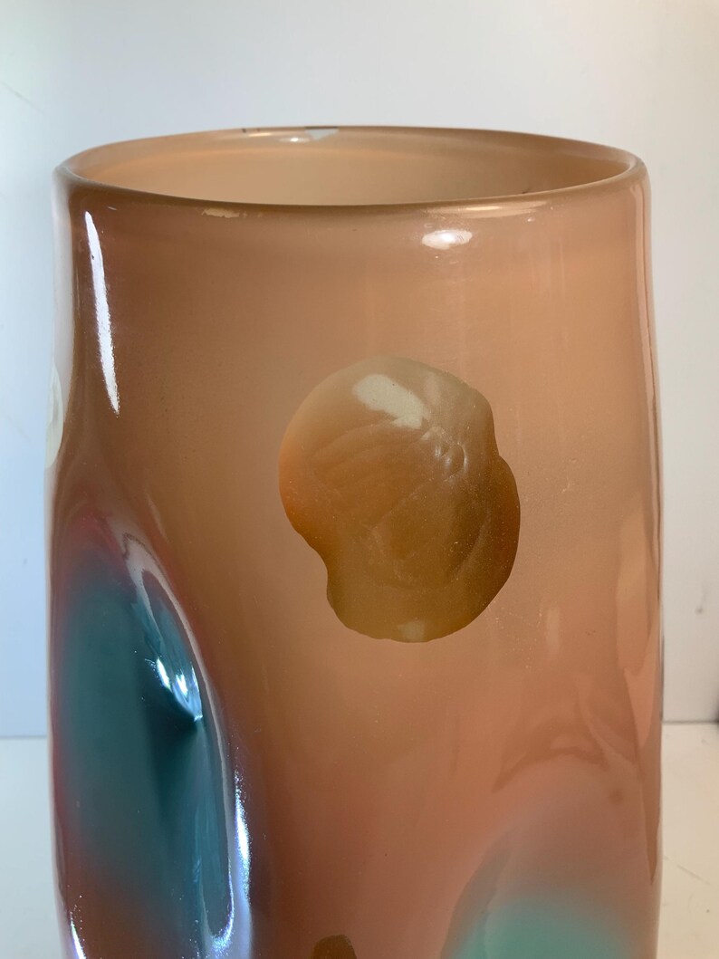 May include: A tall, cylindrical vase with a peach-colored exterior. The glass has a glossy finish with abstract shapes of teal and brown. The vase has a wide opening and a smooth, rounded base. The design is modern and artistic.