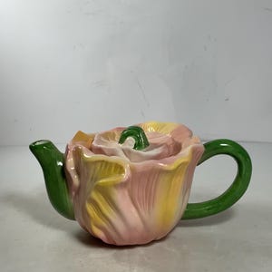 Può includere: Una teiera in ceramica a forma di fiore di tulipano. La teiera ha una combinazione di colori rosa, giallo e verde. Il manico e il beccuccio sono verdi, mentre i petali del fiore sono rosa e gialli.