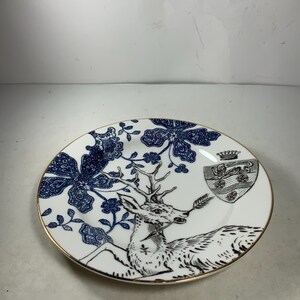 May include: A white porcelain plate with a blue and black floral design and a deer with antlers. The deer is holding a branch in its mouth. There is a crest with a crown on the plate.
