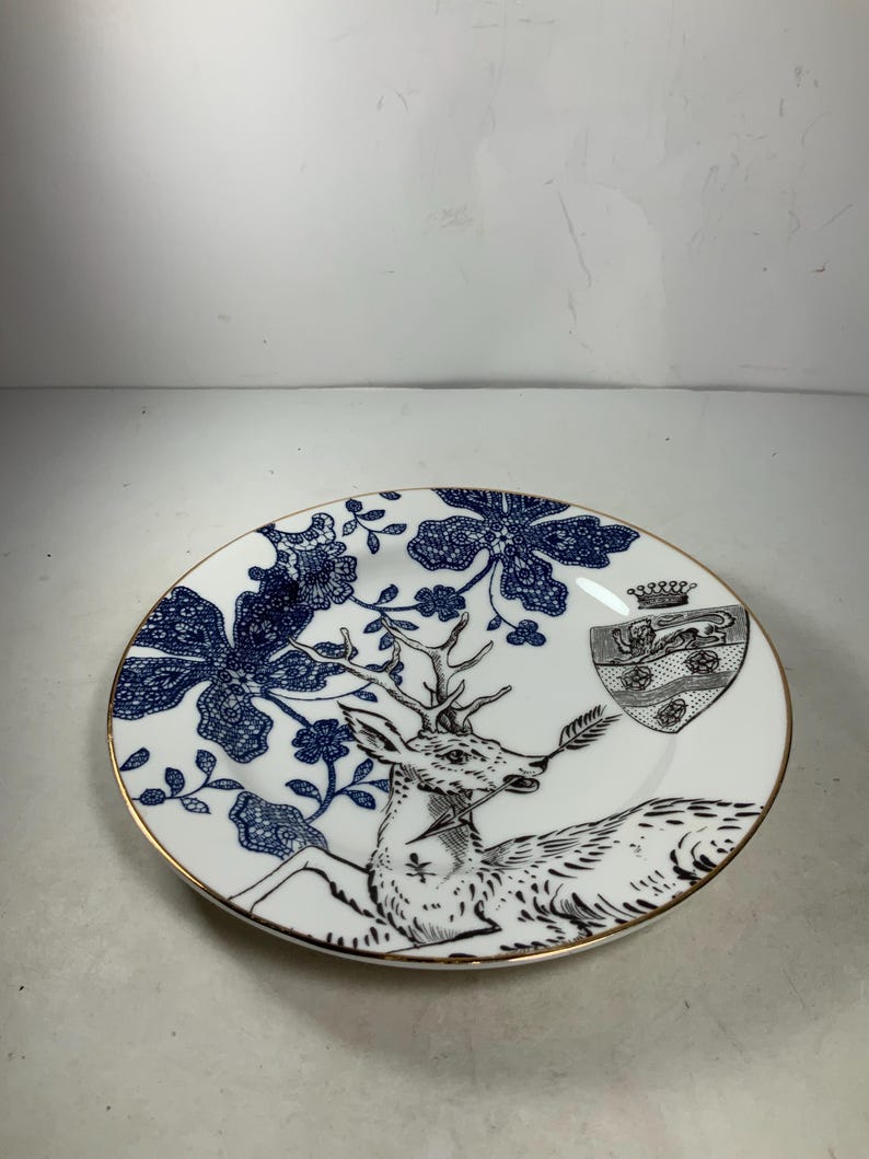 May include: A white porcelain plate with a blue and black floral design and a deer with antlers. The deer is holding a branch in its mouth. There is a crest with a crown on the plate.