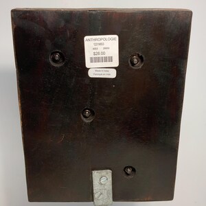 May include: A dark brown, rectangular wooden object with a metal stand. The object has a sticker that reads "ANTHROPOLOGIE" and "Made in India". The object is attached to a silver metal stand with screws.