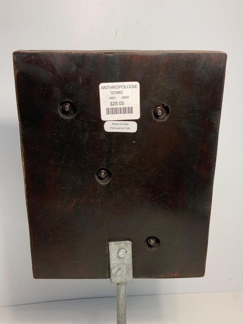 May include: A dark brown, rectangular wooden object with a metal stand. The object has a sticker that reads "ANTHROPOLOGIE" and "Made in India". The object is attached to a silver metal stand with screws.