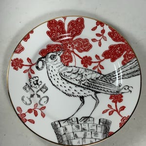 May include: A white porcelain plate with a gold rim features a black and white illustration of a bird perched on a brick chimney. The bird is holding a key in its beak. The background is a red floral pattern.