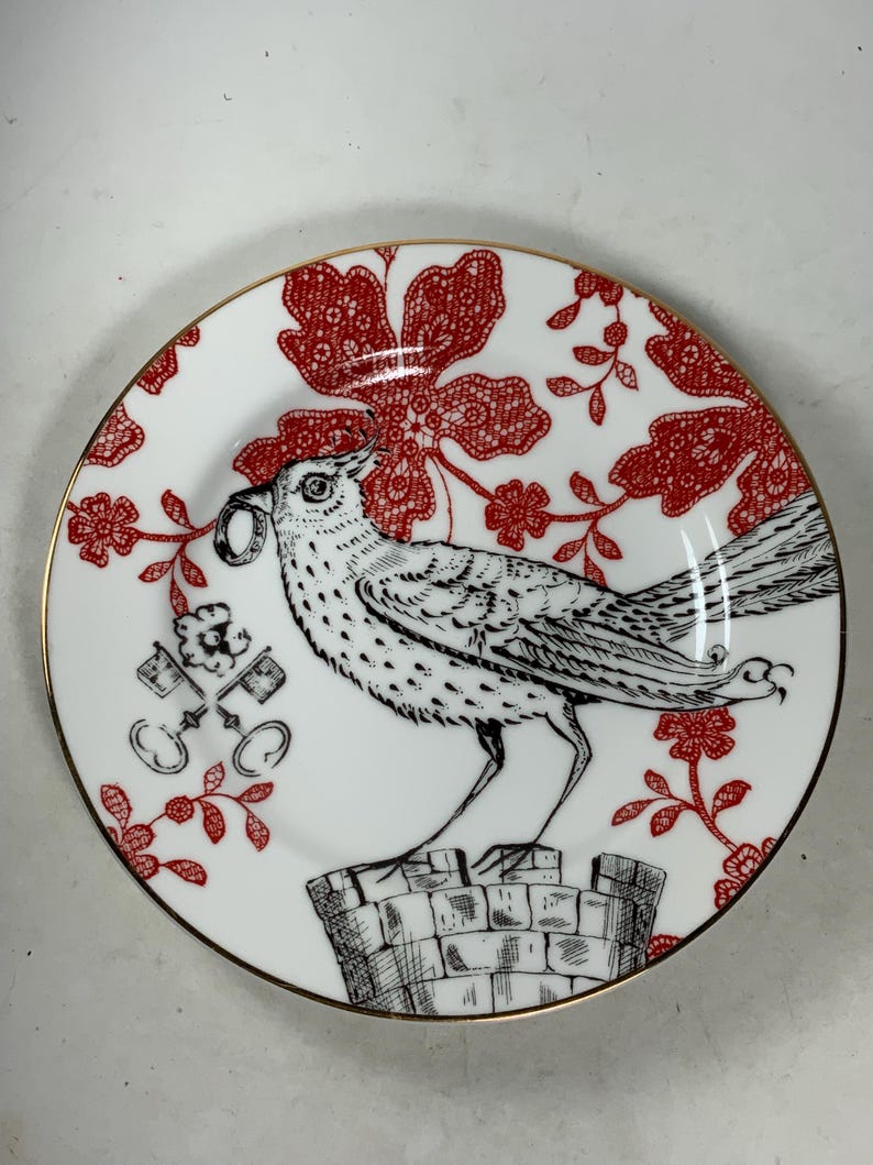 May include: A white porcelain plate with a gold rim features a black and white illustration of a bird perched on a brick chimney. The bird is holding a key in its beak. The background is a red floral pattern.