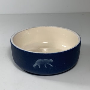 Peut inclure: Petit bol rond en céramique, extérieur bleu marine et intérieur crème. Une silhouette bleu clair d'un ours est représentée sur le côté. Le bol présente un design simple et minimaliste.