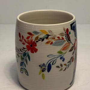 Puede incluir: Una taza de cer&aacute;mica blanca con un dise&ntilde;o floral. La taza cil&iacute;ndrica est&aacute; adornada con flores y hojas coloridas en rojo, azul, amarillo y naranja. El dise&ntilde;o envuelve la taza.