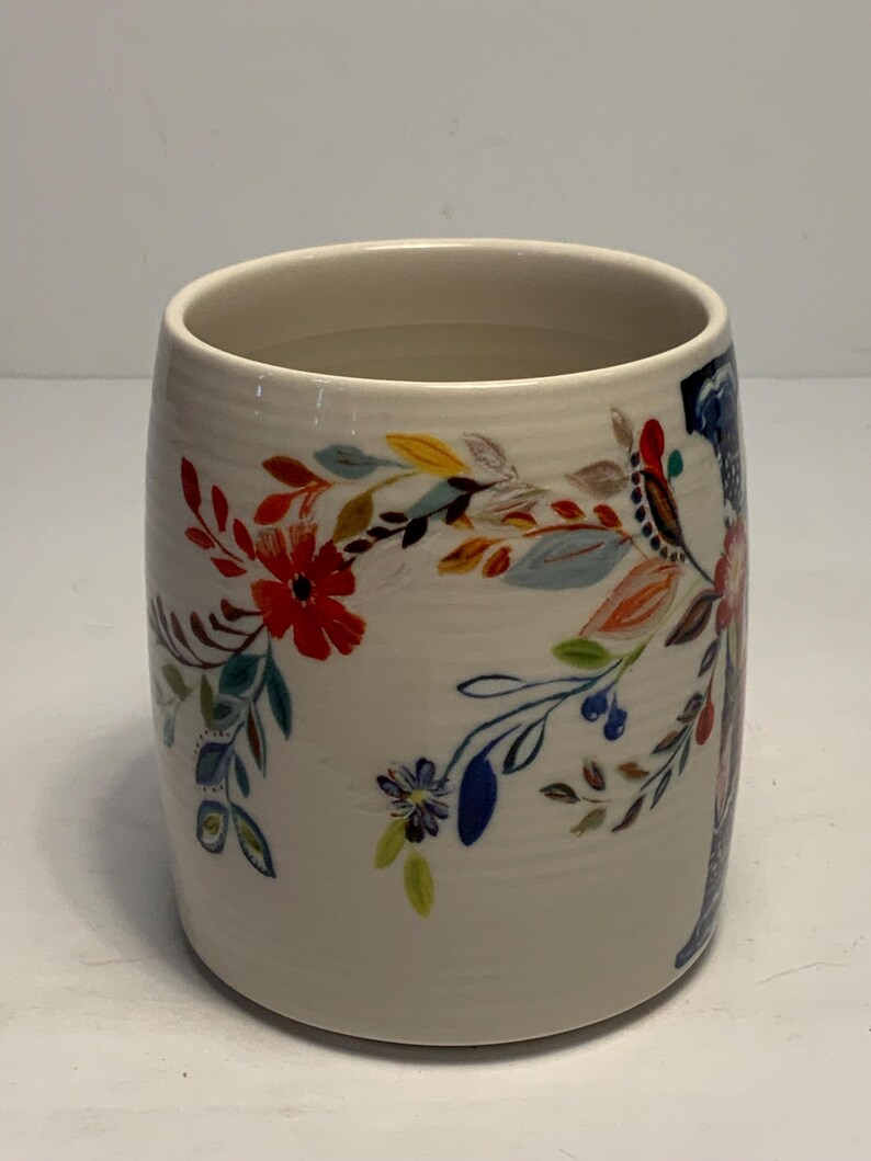 Puede incluir: Una taza de cer&aacute;mica blanca con un dise&ntilde;o floral. La taza cil&iacute;ndrica est&aacute; adornada con flores y hojas coloridas en rojo, azul, amarillo y naranja. El dise&ntilde;o envuelve la taza.