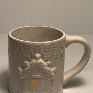 Peut inclure: Une tasse en c&eacute;ramique cr&egrave;me avec une anse. La tasse pr&eacute;sente un motif de maison en pain d'&eacute;pices en relief avec la lettre "C" en or. La tasse est d&eacute;cor&eacute;e de flocons de neige et de motifs de feuilles en relief.
