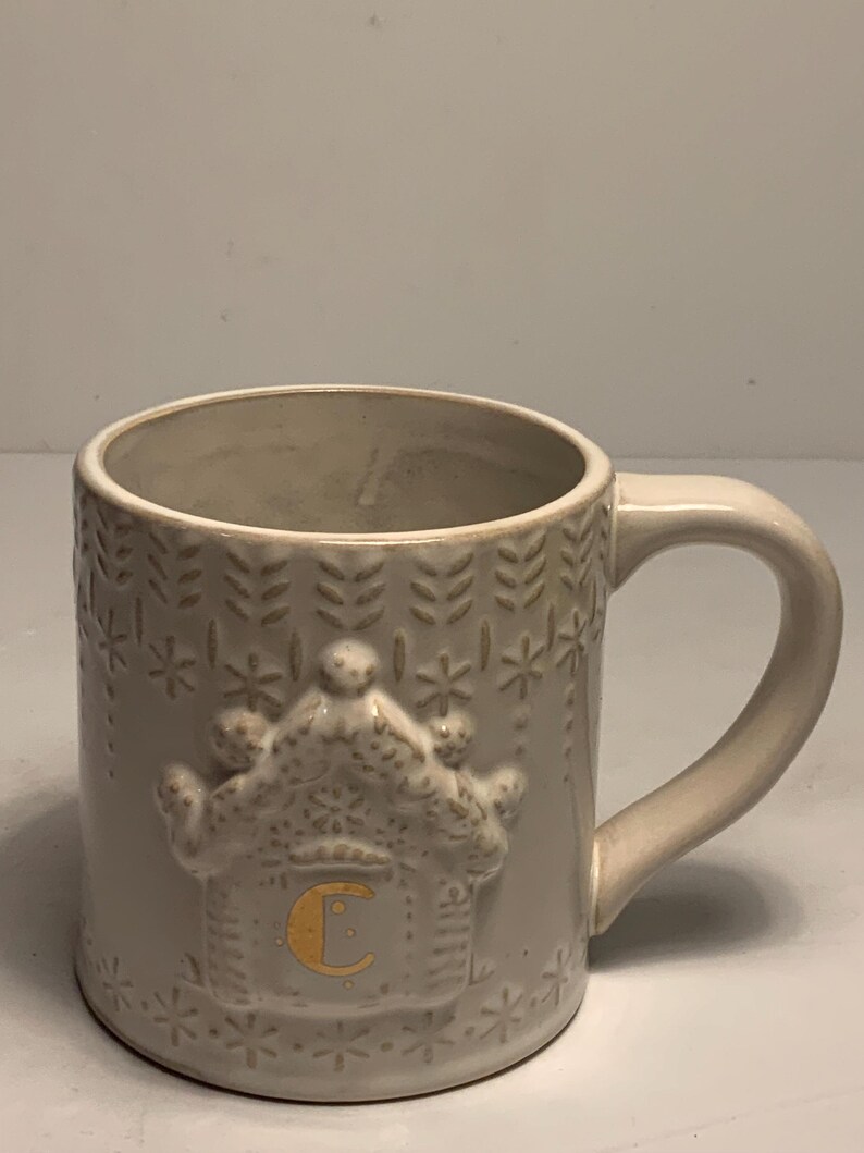 Peut inclure: Une tasse en c&eacute;ramique cr&egrave;me avec une anse. La tasse pr&eacute;sente un motif de maison en pain d'&eacute;pices en relief avec la lettre "C" en or. La tasse est d&eacute;cor&eacute;e de flocons de neige et de motifs de feuilles en relief.