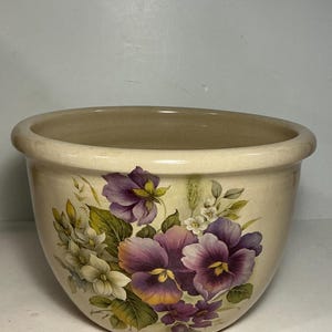May include: A cream-colored ceramic bowl with a wide, rounded rim. The exterior features a detailed floral illustration of purple and white pansies with green leaves and stems, wrapping around the front of the rounded vessel.