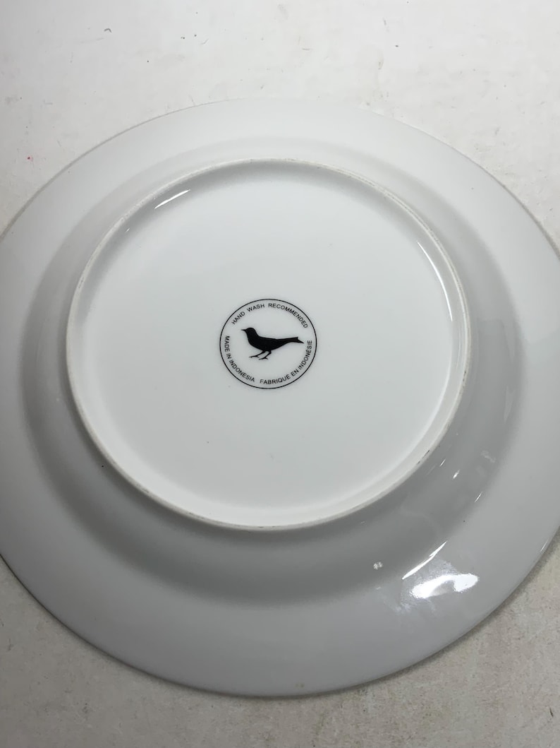 May include: White ceramic plate with a black bird logo on the bottom. The logo reads "Hand Wash Recommended, Made in Indonesia, Fabrique en Indonesie".