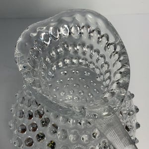 May include: Clear glass pitcher with a textured, hobnail design. The pitcher features a spout and a handle with vertical ridges. The hobnail pattern covers the body of the pitcher, creating a unique, decorative look.