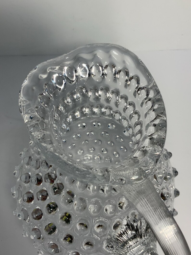 May include: Clear glass pitcher with a textured, hobnail design. The pitcher features a spout and a handle with vertical ridges. The hobnail pattern covers the body of the pitcher, creating a unique, decorative look.