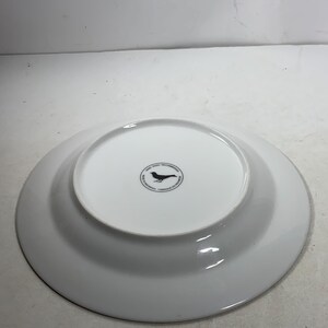 May include: White ceramic dinner plate with a black bird logo on the bottom. The logo reads "Good Ware Recommended, Made in Indonesia, Carbon Neutral"