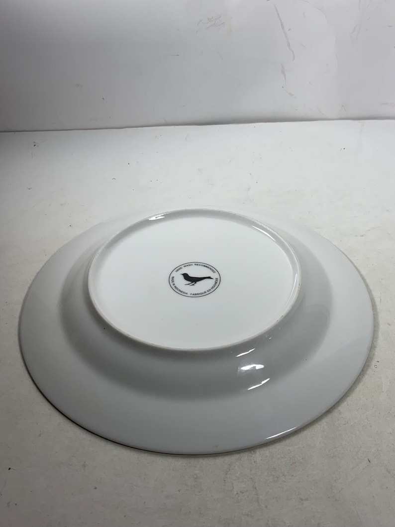 May include: White ceramic dinner plate with a black bird logo on the bottom. The logo reads "Good Ware Recommended, Made in Indonesia, Carbon Neutral"