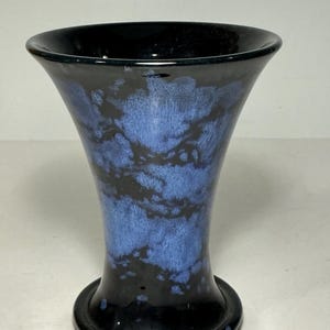 May include: A flared ceramic vase featuring a glossy black base glaze with a mottled, cloud-like pattern of vibrant blue glaze across the centre. The vase has a wide, circular opening at the top and a stable, rounded base, showcasing a smooth finish.