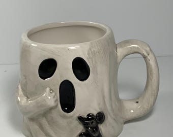 Può includere: Una tazza in ceramica bianca a forma di fantasma con un viso nero e un piccolo topo nero sul lato. La tazza ha un manico ed è perfetta per Halloween.
