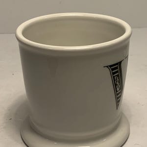 Puede incluir: Un tarro de afeitar de cer&aacute;mica blanca con una base acampanada y un borde ancho. El tarro presenta una letra "V" en blanco y negro en el lateral. El tarro es cil&iacute;ndrico y tiene un acabado brillante.