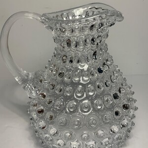 May include: A clear glass pitcher with a textured surface of small, rounded bumps. The pitcher has a curved handle and a spout with a scalloped edge. The overall design is reminiscent of a hobnail pattern, creating a decorative and functional piece.