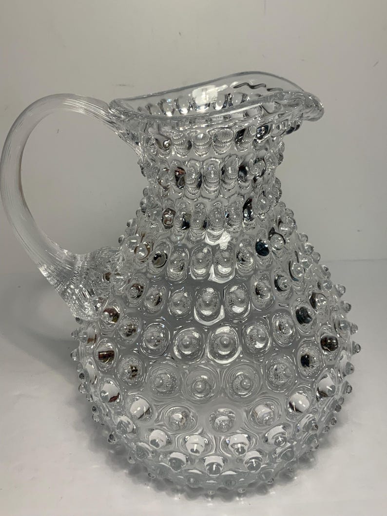 May include: A clear glass pitcher with a textured surface of small, rounded bumps. The pitcher has a curved handle and a spout with a scalloped edge. The overall design is reminiscent of a hobnail pattern, creating a decorative and functional piece.