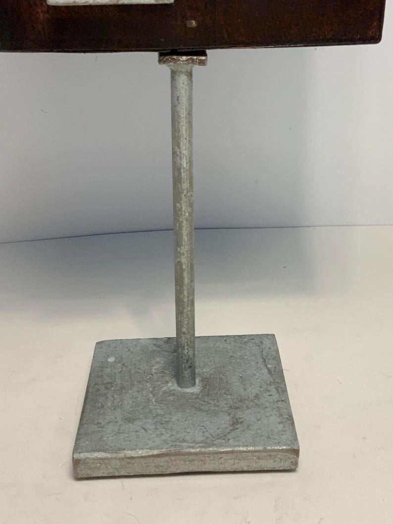 May include: A metal display stand with a square base and a vertical support. The stand is made of a silver-colored metal. The base is a square shape, and the vertical support is a cylindrical rod.