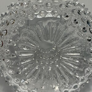 May include: A clear glass bowl with a textured surface. The bowl has a pattern of small, raised circles around the rim and a starburst design in the center. The bowl is likely used for serving or display.