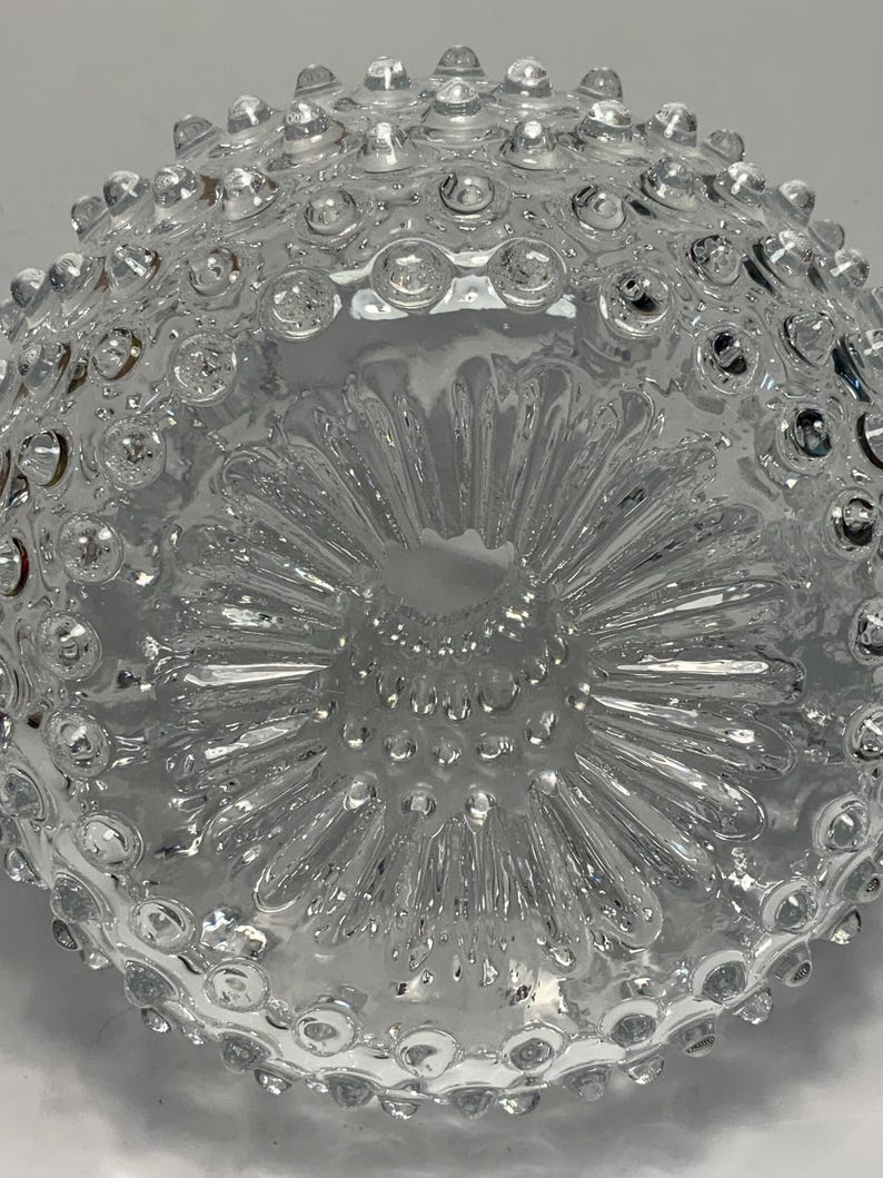 May include: A clear glass bowl with a textured surface. The bowl has a pattern of small, raised circles around the rim and a starburst design in the center. The bowl is likely used for serving or display.