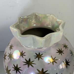 May include: A pearlescent white ceramic vase with a star pattern. The vase has a round body and a ruffled, star-shaped rim. Gold stars are scattered across the surface. The vase is suitable for home decor.