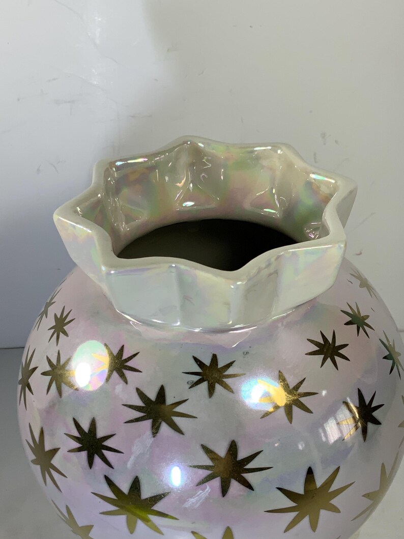 May include: A pearlescent white ceramic vase with a star pattern. The vase has a round body and a ruffled, star-shaped rim. Gold stars are scattered across the surface. The vase is suitable for home decor.