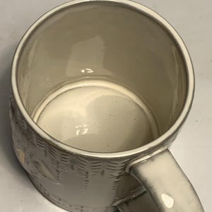 May include: A cream-colored ceramic mug with a textured basket weave design. The mug has a rounded handle and a smooth, glossy interior. The mug is empty and appears to be clean.