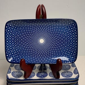 Op de afbeelding: Een stapel rechthoekige keramische serveerschalen. De bovenste schaal heeft een blauwe achtergrond met een wit geometrisch patroon. De onderste schalen hebben een witte achtergrond met een blauw cirkelvormig patroon. De schalen staan op een houten standaard.