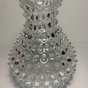 May include: Clear glass pitcher with a textured, bubble-like surface. The pitcher has a handle and a spout, and the body is covered in small, rounded bumps. The overall design is vintage and decorative, suitable for serving beverages.