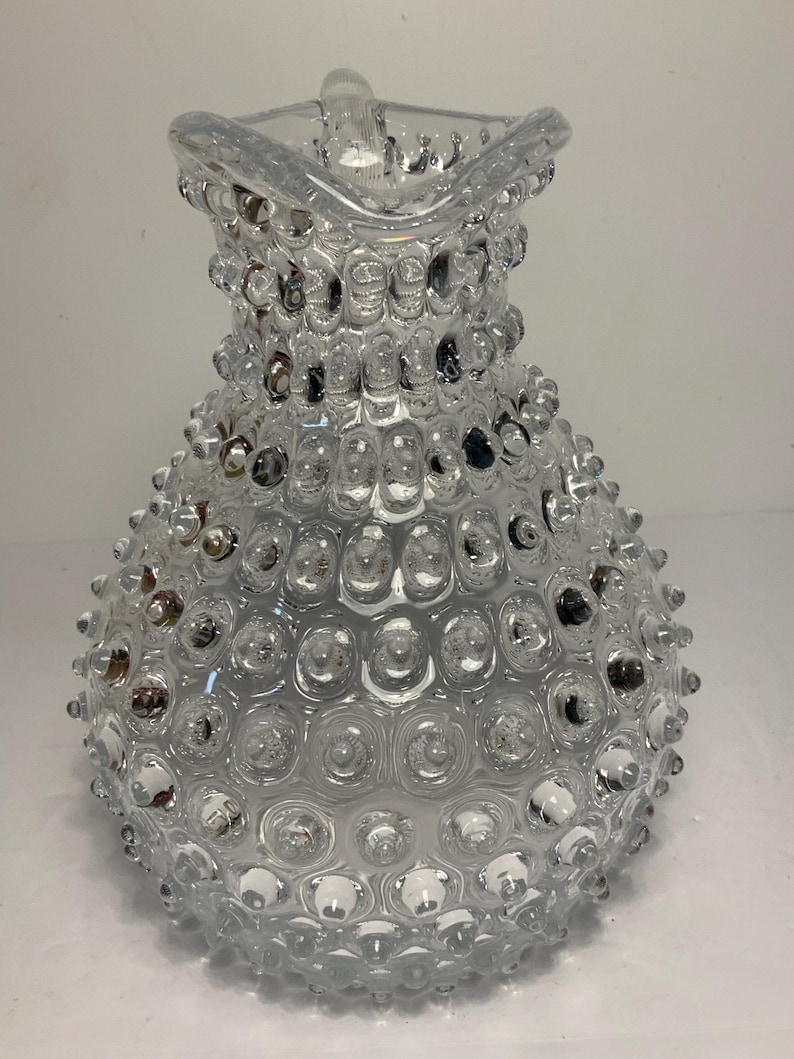 May include: Clear glass pitcher with a textured, bubble-like surface. The pitcher has a handle and a spout, and the body is covered in small, rounded bumps. The overall design is vintage and decorative, suitable for serving beverages.