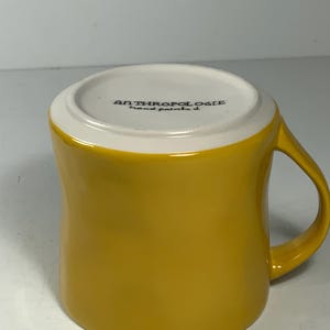 Puede incluir: Una taza de cer&aacute;mica amarilla mostaza con un asa curvada. El borde interior es blanco y la base de la taza tiene el texto "ANTHROPOLOGIE hand painted". La taza tiene un acabado brillante y est&aacute; dise&ntilde;ada para bebidas calientes.