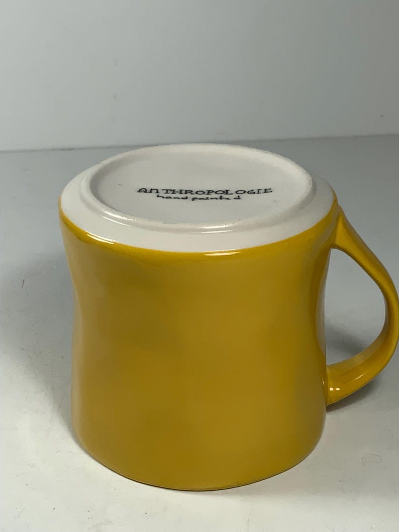 Puede incluir: Una taza de cer&aacute;mica amarilla mostaza con un asa curvada. El borde interior es blanco y la base de la taza tiene el texto "ANTHROPOLOGIE hand painted". La taza tiene un acabado brillante y est&aacute; dise&ntilde;ada para bebidas calientes.