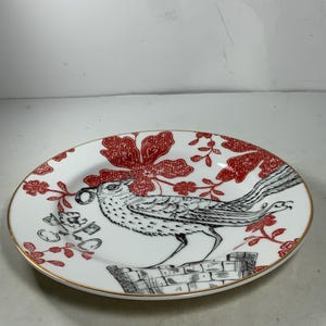 May include: A white ceramic plate with a red and black bird design. The bird is perched on a stack of books and surrounded by red floral patterns.