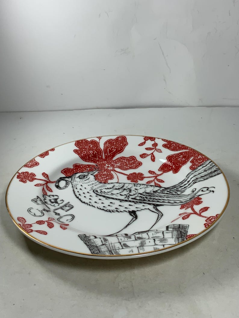 May include: A white ceramic plate with a red and black bird design. The bird is perched on a stack of books and surrounded by red floral patterns.