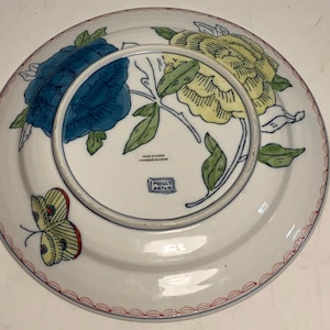 May include: White ceramic plate with a floral design featuring blue and yellow flowers, green leaves, and a yellow butterfly with red accents. The plate has a red and white border and the text "MADE IN CHINA" and "MOLLY HATCH".