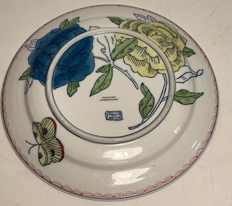 May include: White ceramic plate with a floral design featuring blue and yellow flowers, green leaves, and a yellow butterfly with red accents. The plate has a red and white border and the text "MADE IN CHINA" and "MOLLY HATCH".