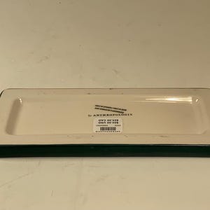 May include: Rectangular ceramic tray with a cream-colored interior and a dark green border. The tray has a slightly raised edge and a label that reads "by Anthropologie".